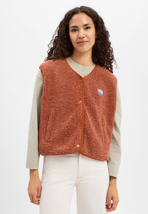 Vrouw met krullend haar die een roestkleurig gestructureerd vest draagt over een licht shirt met lange mouwen en witte broek, staand tegen een effen achtergrond.