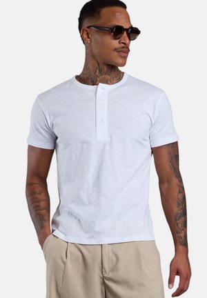 Man with neck and arm tattoos wearing dark sunglasses, a white short-sleeve henley shirt, and beige pants with hands in pockets.