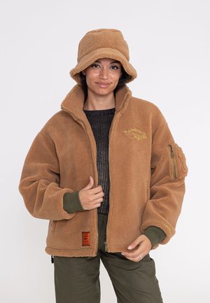 Frau trägt eine braune Fleecejacke und passenden Fischerhut, dazu olivgrüne Hose und einen dunklen Strickpullover darunter.