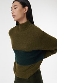 Oversized trui in olijfgroen met een teal horizontale streep. Gemaakt van zacht materiaal met een geribde col en verlengde schouders.
