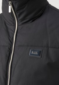 Sort pufferjakke med høj krave, sølvfarvet lynlås og et logo-patch. Glat stof med en mat finish og vandret syningsdetalje.
