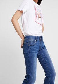 T-shirt blanc avec un motif graphique, associé à un jean denim bleu. Le jean a une coupe slim et des détails subtils d'usure.