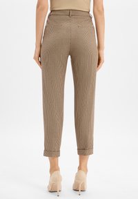 Femme portant un pantalon à carreaux beige longueur cheville et des talons hauts beiges, se tenant debout avec les mains détendues de chaque côté.