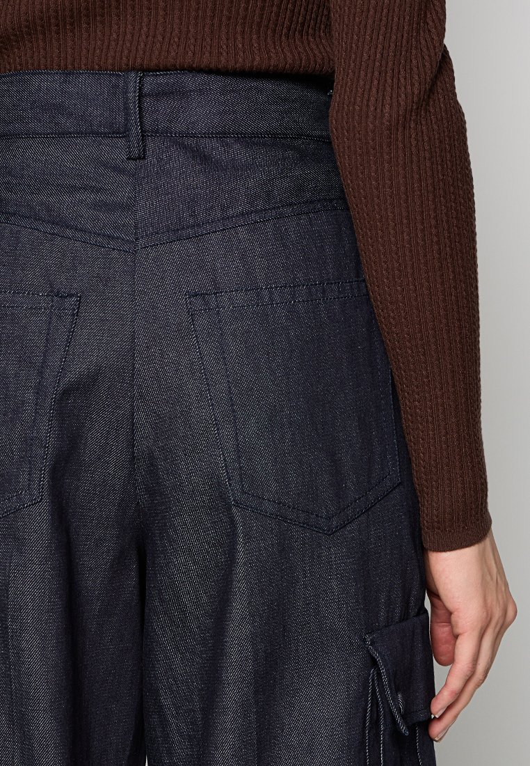 Donkere spijker cargo pants met twee achterzakken, zichtbaar stiksel en een relaxte pasvorm. Een geribbelde bruine top die gedeeltelijk zichtbaar is aan de rechterkant.
