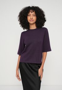 Violets īsroku džemperis ar apaļu kakla izgriezumu un vertikālu ribēto dizainu. Valkāts kopā ar gludu melnu svārku.