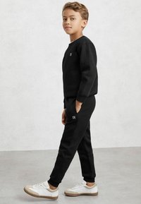 Hanorac negru cu mâneci lungi și pantaloni de jogging asortați, amândouă cu un finisaj texturat și un mic logo alb, asortate cu adidași albi și bej.