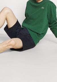 Zelená bavlněná mikina s kulatým výstřihem a logem, doplněná tmavě modrými šortkami, nabízí volný střih a jemnou texturu.