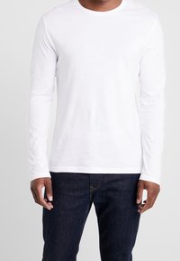 Langärmliges weißes Baumwoll-T-Shirt mit Rundhalsausschnitt, kombiniert mit dunkelblauen Denim-Jeans. Glatte Textur, keine sichtbaren Muster oder Akzente.