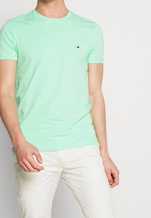 Man draagt een muntgroen T-shirt met korte mouwen en crèmekleurige broek, staand tegen een effen lichte achtergrond.