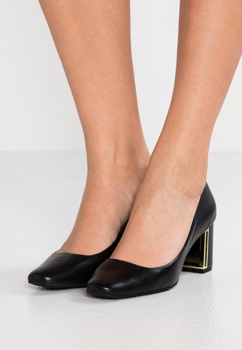 Pieds portant des chaussures en cuir noir à talon bas bloc avec un détail métallique doré sur le talon, sur un fond clair uni.