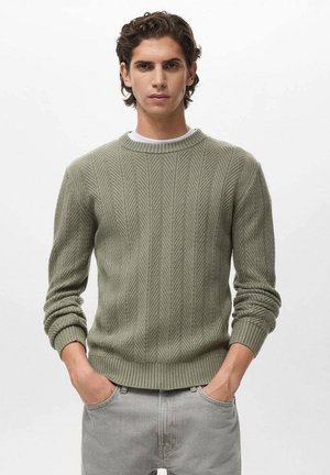 Suéter de punto verde oliva con un patrón de espina de pescado texturizado, cuello redondo, puños y dobladillo acanalados. Combinado con pantalones gris claro.