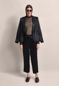 Schwarze Satin-Blazer über einem transparenten, bronzefarbenen Strickoberteil, kombiniert mit schwarzen weit geschnittenen Hosen und schwarzen Slippern. Klare Linien, minimalistisches Design.