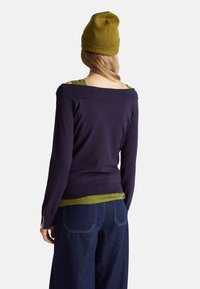 Pulovr v námořnické modři s odkrytými rameny, žebrovaným výstřihem, navlečený přes zelenou blůzu. Spárováno s džínovými kalhotami s vysokým pasem.