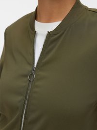 Veste bomber vert olive avec fermeture à zip, col côtelé et texture lisse. Superposée sur une chemise blanche.