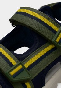 Κοντινό πλάνο σε λουριά σανδαλιού πράσινου και ναυτικού μπλε με κίτρινες και σκούρες μπλε ρίγες και πράσινες πλαστικές πόρπες.