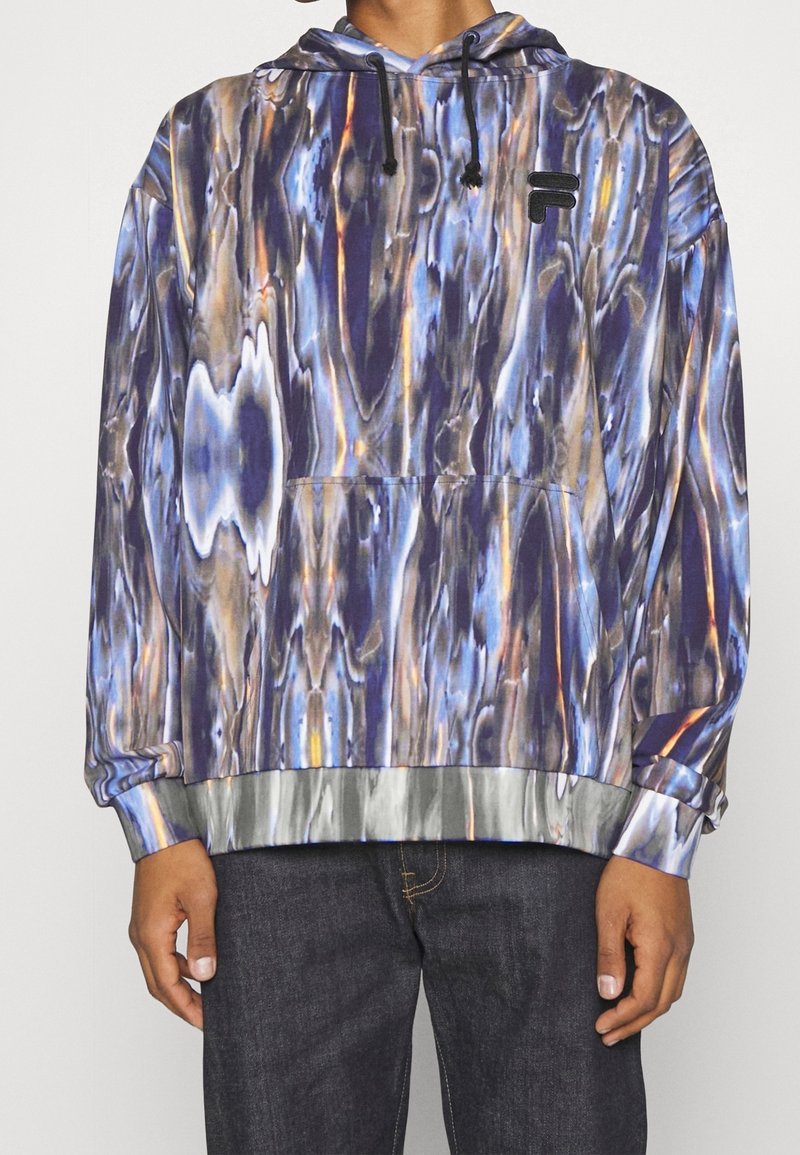 Personne portant un sweat à capuche à motifs bleus, violets et beige avec un logo noir sur la poitrine gauche et un jean foncé.