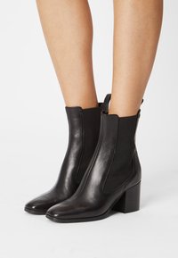 Piernas que llevan elegantes botas de cuero negro con tacones de bloque y paneles laterales elásticos sobre un fondo blanco.