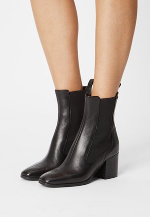 Legs wearing sleek black leather ankle boots with block heels and elastic side panels on a white background.