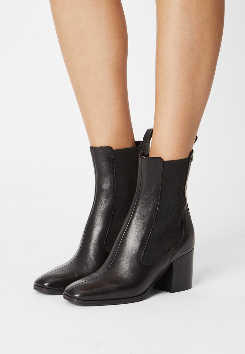 Piernas que llevan elegantes botas de cuero negro con tacones de bloque y paneles laterales elásticos sobre un fondo blanco.