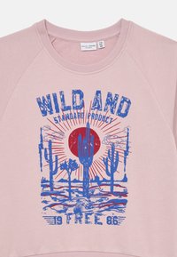 Rosa genser med blå grafisk trykk av kaktuser, sol og teksten "WILD AND STANDARD PRODUCT" samt "19 FREE 86."