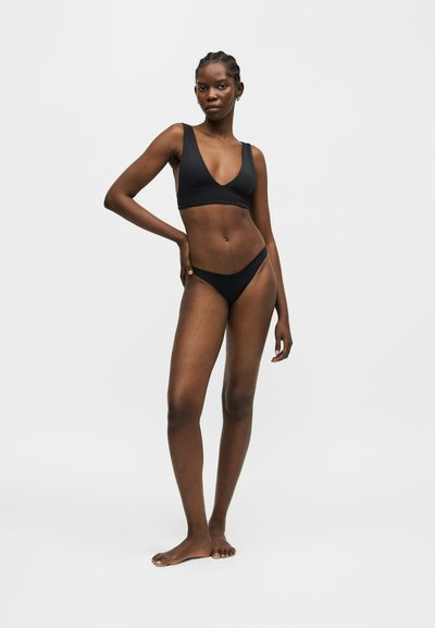 Femme debout pieds nus portant un bikini noir avec un haut à col en V profond et un bas assorti sur un fond clair uni.