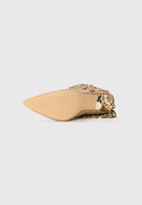 Puntige beige suede schoen met een lage gouden blokhak, versierd met franjes en kleine rhinestones, zool naar boven op een witte achtergrond.
