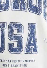 Tissu blanc avec un grand texte bleu vieilli indiquant "FIER USA" et un texte plus petit en dessous. Design simple et décontracté.