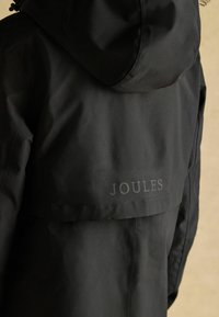 Jachetă impermeabilă neagră cu glugă și clapetă pe spate, având logo-ul "JOULES" într-un font subtil gri. Textură exterioară netedă, fără modele vizibile.