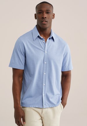 Homme portant une chemise bleu clair à manches courtes boutonnée et un pantalon beige, debout, une main dans la poche sur un fond uni.