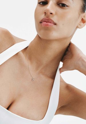 Collar de plata con un pequeño colgante, llevado sobre piel desnuda, junto a una blusa blanca sin mangas, resaltando el diseño delicado de la cadena.