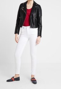 Giacca da motociclista nera in pelle con spalle trapuntate, t-shirt rossa, jeans skinny bianchi e mocassini blu con dettagli rossi e catena.
