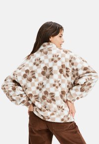 Veste en polaire douce avec des motifs à carreaux et floraux marron sur fond crème, dotée d'un col haut et d'une coupe oversize.