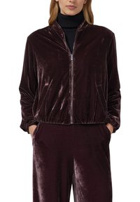 Sieviete, tērpusies tumši purpura zīda jakā ar rāvējslēdzēju un atbilstošās biksēs, rokas ievietotas kabatās, zem melnās turtleneck blūzes.