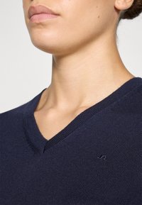 Mörkblå stickad tröja med V-ringning och subtil textur. Har en liten broderad logo nära halsringningen för detaljer.