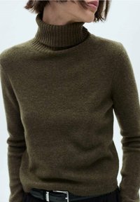 Pull col roulé vert olive en tissu tricoté doux, avec un col côtelé et des manches longues, associé à une ceinture noire.