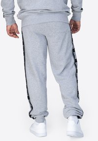 Pantalones de chándal grises con rayas laterales negras que presentan una marca de logotipo, puños elásticos y un ajuste relajado. El material parece suave y con textura.