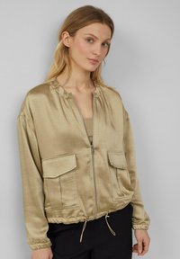 Frau mit hellbraunem Haar, die eine beige satinierte Zip-Jacke mit großen Fronttaschen und schwarze Hosen vor einem schlichten grauen Hintergrund trägt.