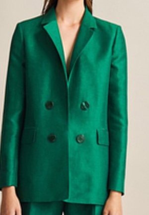 Vrouw die een donkergroene, dubbelgeknoopte blazer met zwarte knopen en bijpassende broek draagt tegen een effen achtergrond.