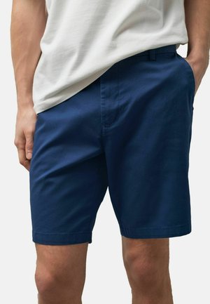 Mann trägt marineblaue knielange Shorts und ein weißes T-Shirt, die Hände in den Taschen, vor einem schlichten Hintergrund.