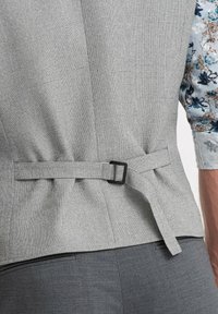 Gray textured vest featuring a back strap with a black metal buckle, paired with dark gray pants. Floral-patterned shirt partially visible.