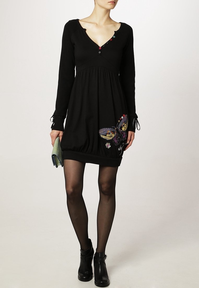 Femme portant une robe noire à manches longues avec une broderie de papillon sur la jupe, des bottines noires, des collants noirs transparents, et tenant une pochette verte.