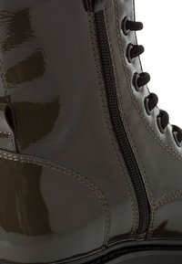 Botine din piele patentă maro, cu fermoar lateral, vârf rotund și șireturi negre contrastante. Textură netedă, cu detalii de cusătură vizibile.