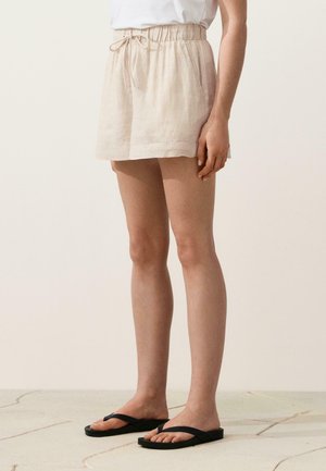 Femme portant un short beige à cordon, un haut blanc et des tongs noires, debout sur un sol carrelé clair devant un fond uni.