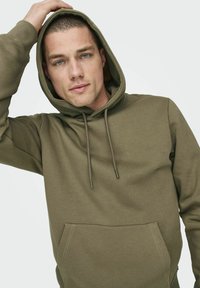 Sudadera verde oliva hecha de tejido suave, con un bolsillo canguro, capucha con cordón y puños acanalados. Textura suave y diseño minimalista.