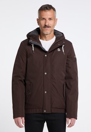 Ein Mann trägt eine dunkelbraune Jacke mit Kapuze und weißen Kordeln, steht vor einem weißen Hintergrund und blickt geradeaus.