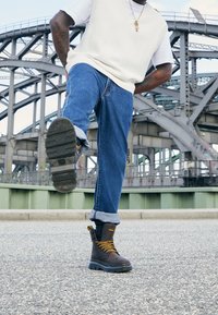 Botas marrones con cordones y suelas negras gruesas, combinadas con jeans azules remangados y una blusa de punto de color claro.