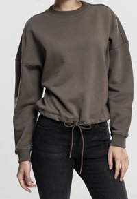 Sudadera marrón de tejido suave, con mangas largas, cuello redondo y un dobladillo con cordón. Combinada con jeans oscuros.