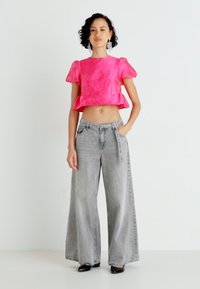 Rozā teksturēts cropped tops ar pūkainām piedurknēm, kombinēts ar pelēkiem augstās jostasvietas, plaša kāju džinsiem. Modelis valkā melnus apavus un minimālus aksesuārus.