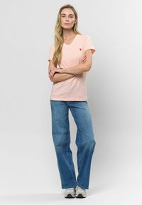 Gaiši rozā v-veida kakla T-krekls, kas izgatavots no kokvilnas, apvienots ar platām ziliem džinsiem. Uz krekla ir neliels sarkans logo. Ikdienas piegriezums un dizains.