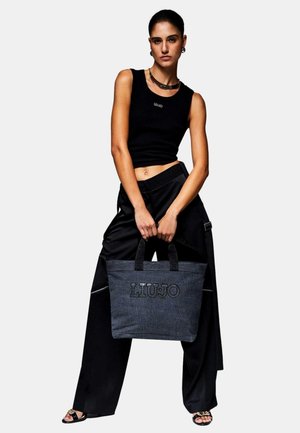 Donna con top crop nero senza maniche e pantaloni a gamba larga tiene una grande borsa tote grigia con la scritta "LIU JO", in piedi davanti a uno sfondo semplice.